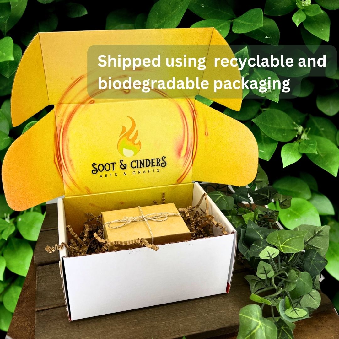 A white box with a yellow interior and lid, containing a wrapped package and brown packing material. The box is labeled 'Soot & Cinders ARTS & CRAFTS' and states 'Shipped using recyclable and biodegradable packaging.'