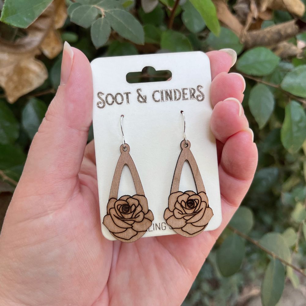 Wooden rose earrings on a 'Soot & Cinders' card held in a hand with a green leafy background.