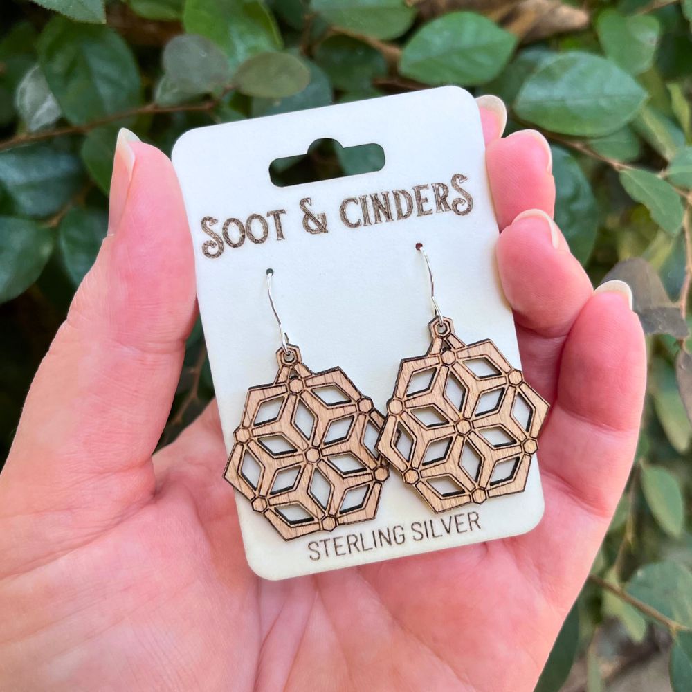 Hand holding a card with geometric earrings labeled 'Soot & Cinders' against a leafy background. They are hexagonal wooden earrings made of red oak and recycled sterling silver that have crisscrossing lines that form many congruent diamonds