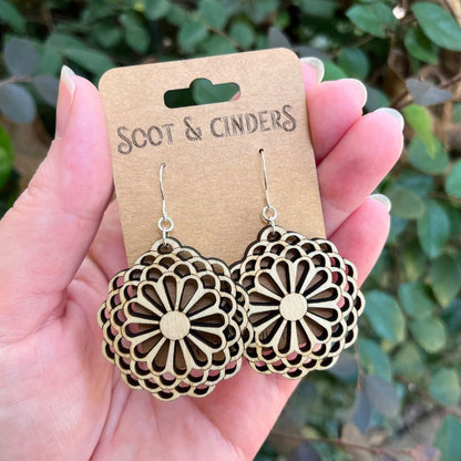 Wooden floral earrings on a 'Soot & Cinders' card held in a hand with a green leafy background.