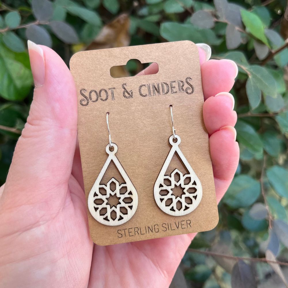 White/beige wooden earrings with silver ear hooks on a 'Soot & Cinders' card held by a hand with a green leafy background