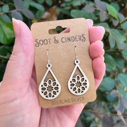 White/beige wooden earrings with silver ear hooks on a 'Soot & Cinders' card held by a hand with a green leafy background
