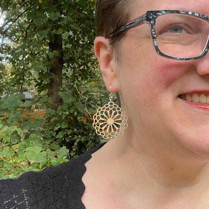 Close-up of a person wearing large wood floral earrings with a green outdoor background