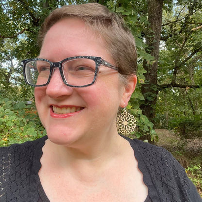 Woman wearing glasses and a black sweater with a forest background wearing large statement earrings in a stylized flower pattern.