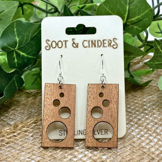 Rectangular solid cherry wood earrings with circle cutouts and recycled sterling silver. Handmade and sustainable. Earrings are on a white card with Soot & Cinders on it and leaves in the background.