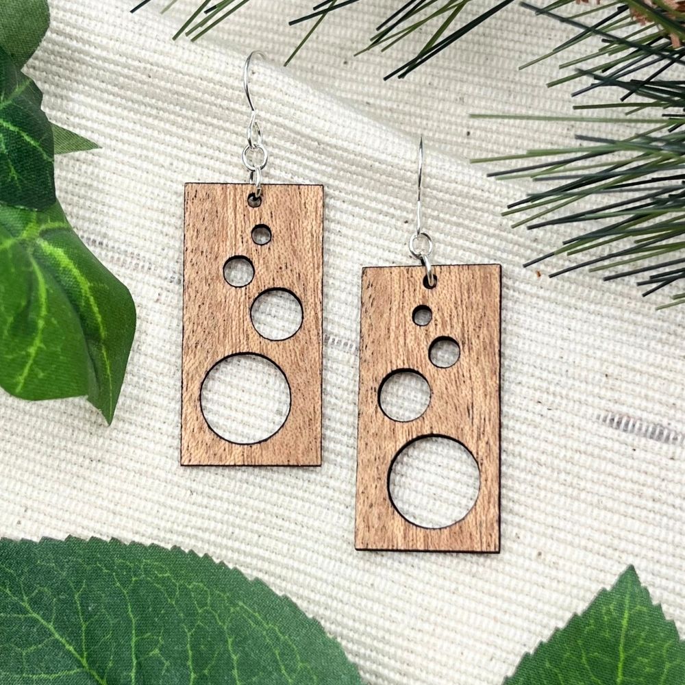 Wooden earrings with circular cutouts on a textured surface with green leaves.