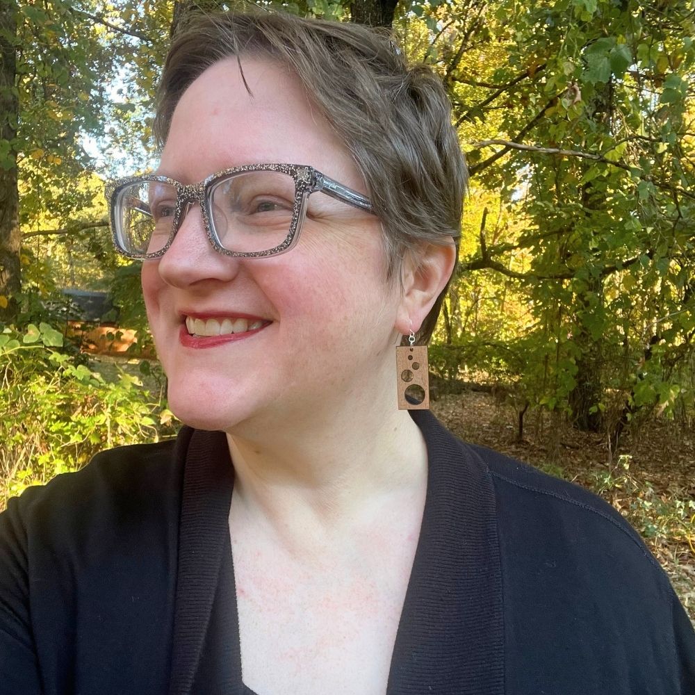Woman with glasses and short hair standing in a forest and wearing rectangular wooden earrings with circular cutouts