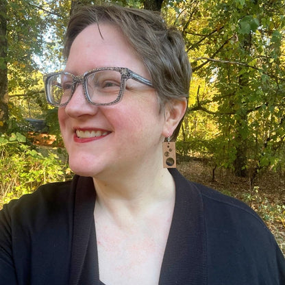 Woman with glasses and short hair standing in a forest and wearing rectangular wooden earrings with circular cutouts