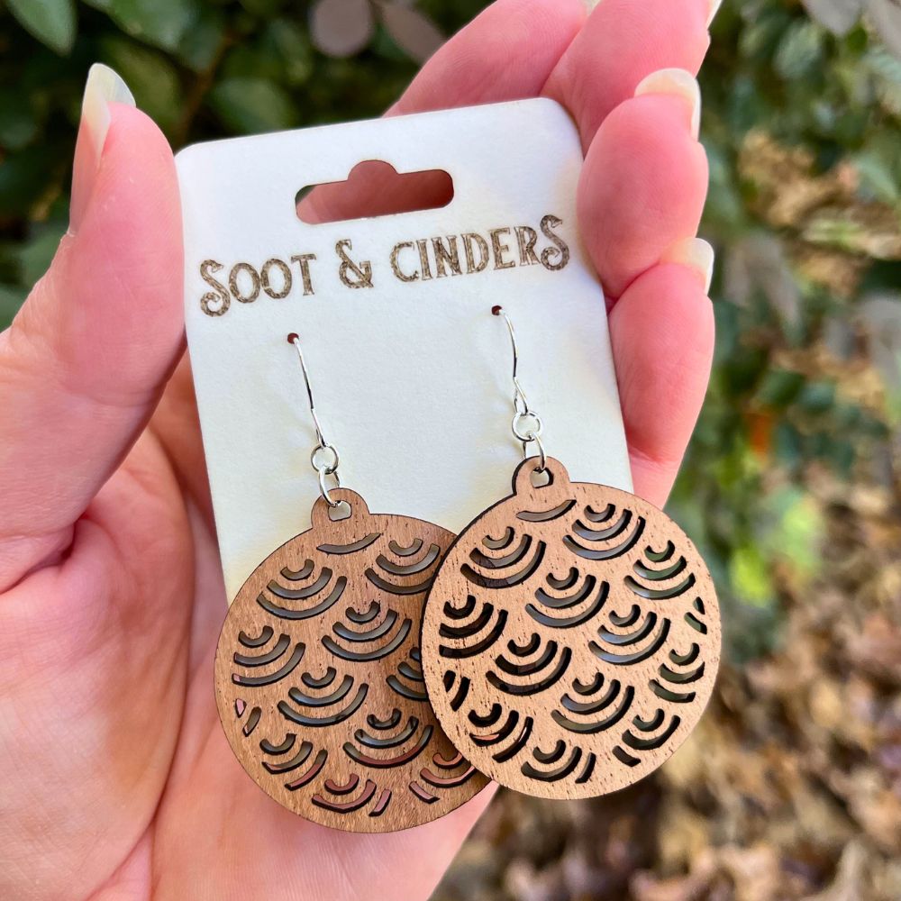 Wooden earrings with intricate patterns held by a hand on a 'Soot & Cinders' branded card.