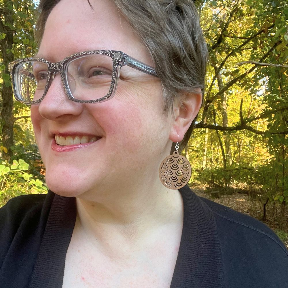 Woman wearing glasses and a black top with cherry wood statement earrings in front of  a forest background