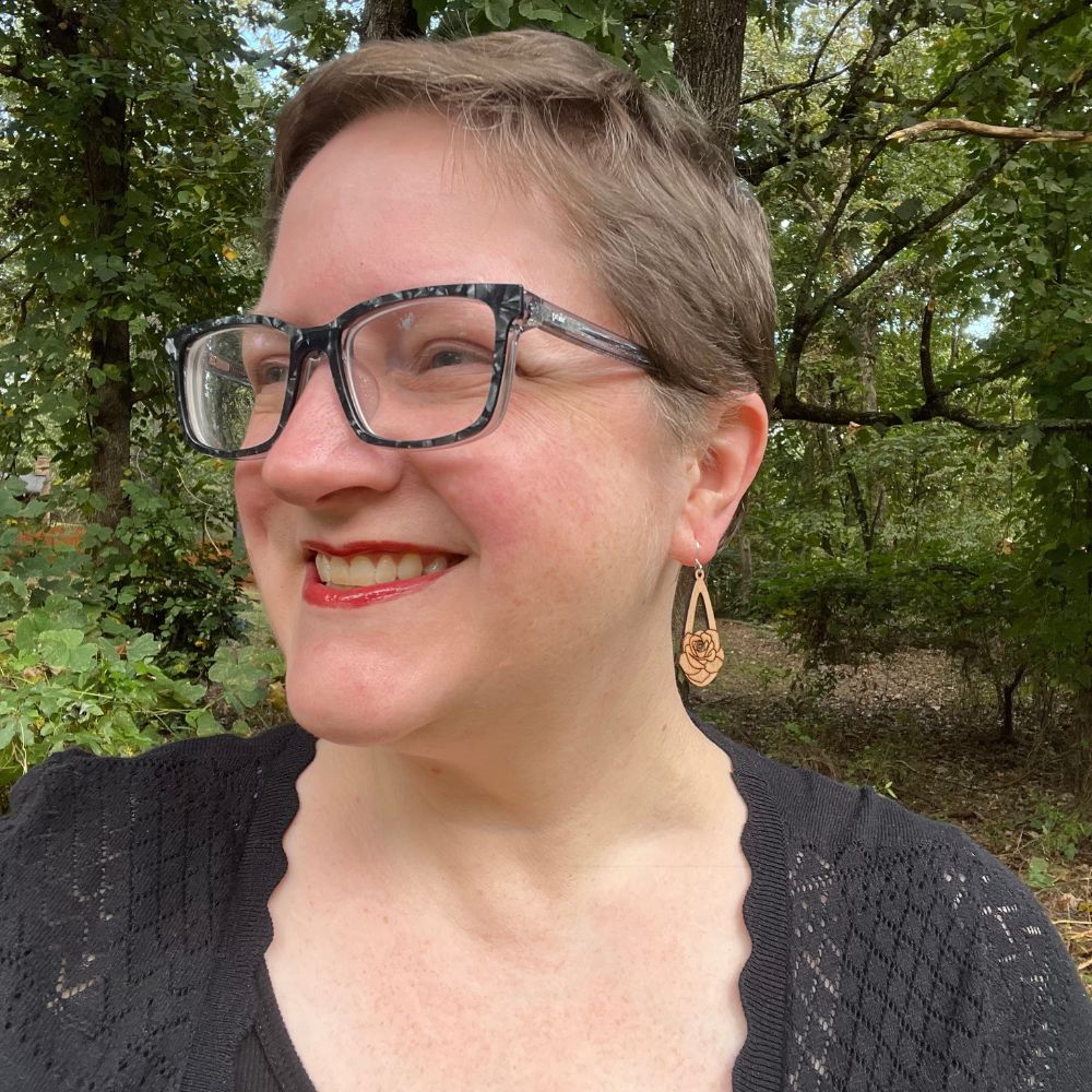 Person with glasses and a black knit cardigan standing outdoors with trees in the background and wearing teardrop-shaped brown wood earring with a rose engraved on them