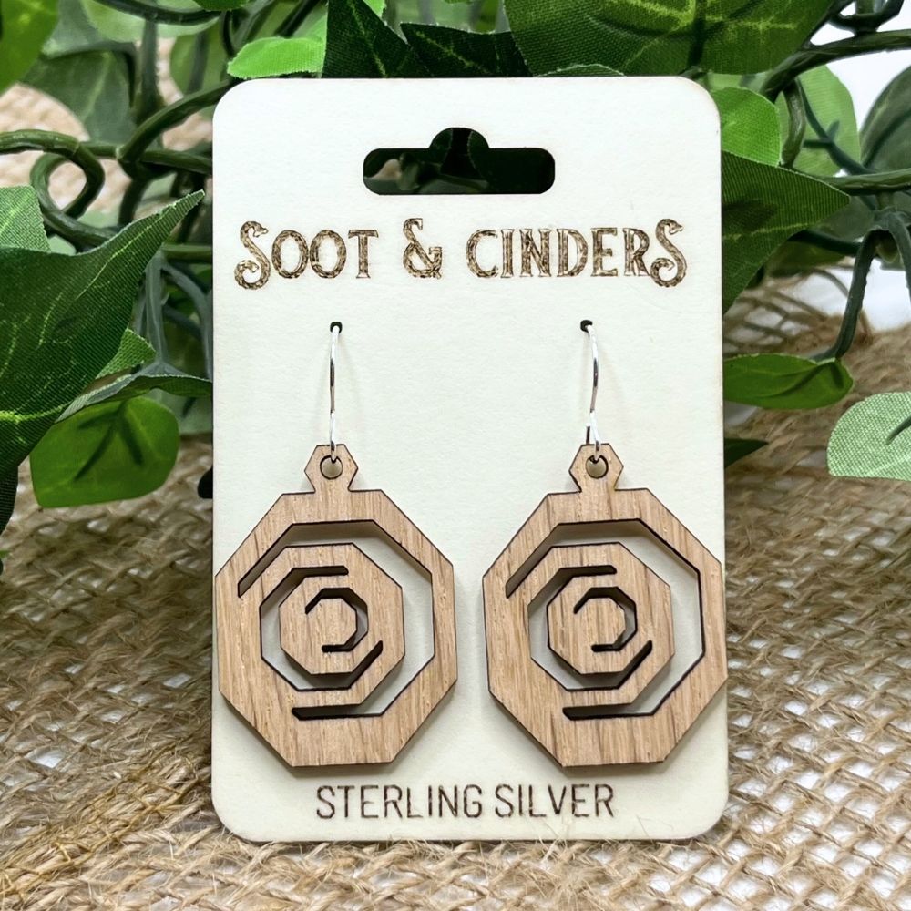Octagonal geometric earrings made of solid red oak wood and recycled sterling silver on a 'Soot & Cinders' branded card with green leaves in the background