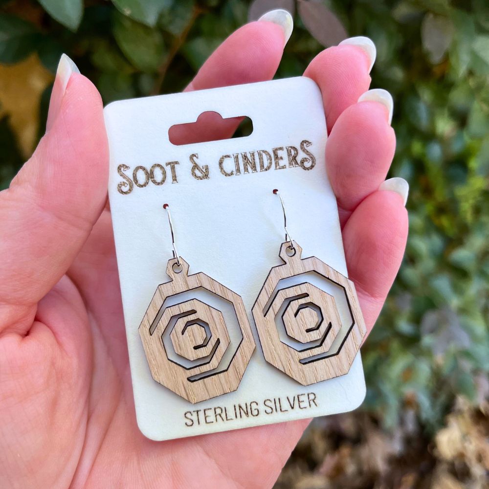 Hand holding a card with wooden earrings labeled 'Soot & Cinders' against a blurred natural background.