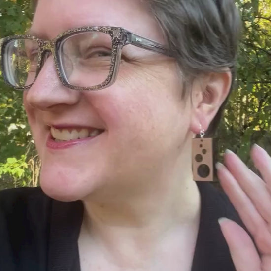 Video showing a woman modeling rectangular cherry wood and sterling silver earrings, then the earrings are shown lying in a woman's hand while text about the earrings' sustainability is seen. The video ends with the Soot & Cinders logo.