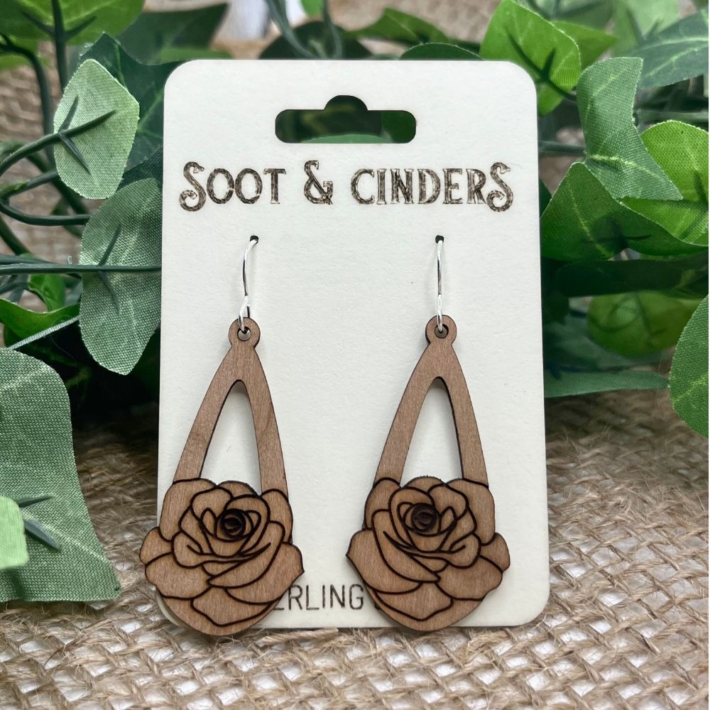 Wooden rose-shaped earrings on a 'Soot & Cinders' branded card with green leaves in the background.