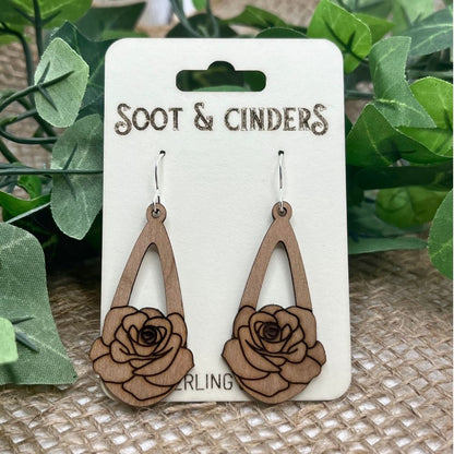 Wooden rose-shaped earrings on a 'Soot & Cinders' branded card with green leaves in the background.
