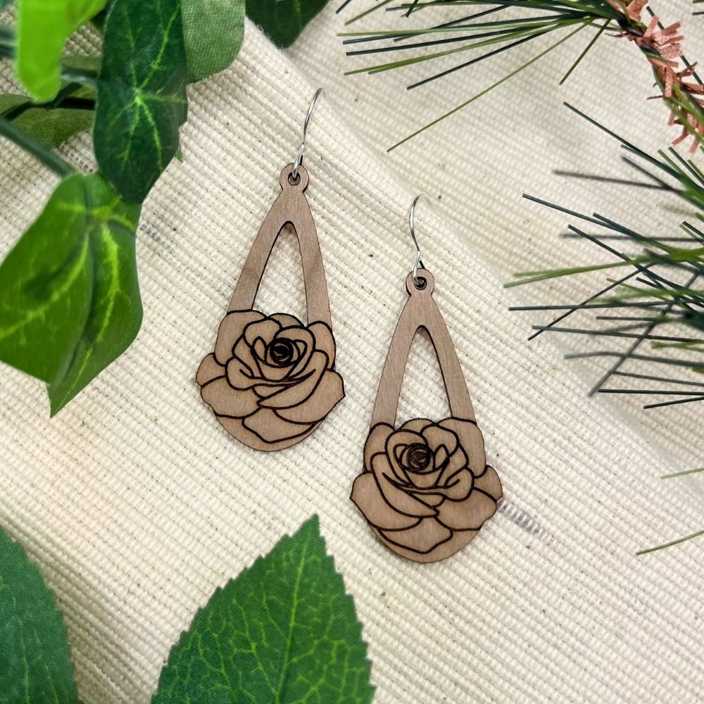 mahogany teardrop earrings with a rose engrave on them rest on a textured fabric background surrounded by leaves