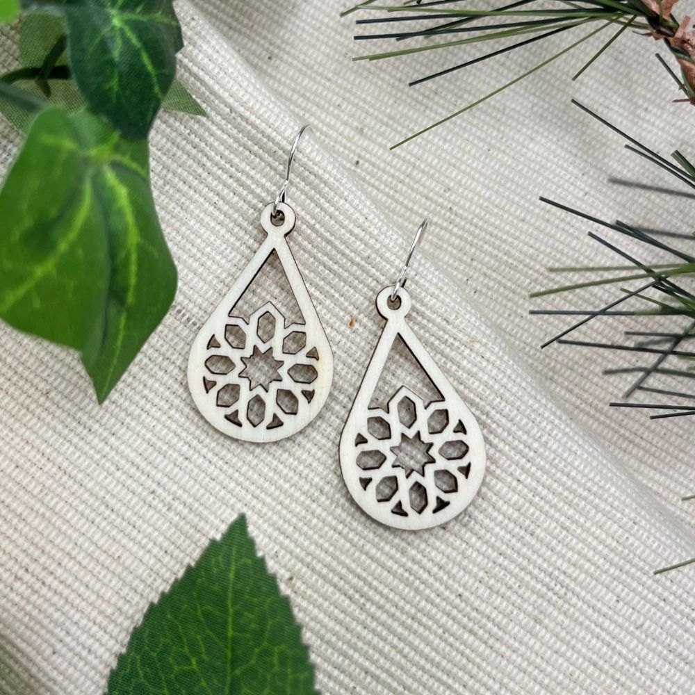 Sterling silver and white wood teardrop earrings with intricate designs on a textured surface with green leaves.