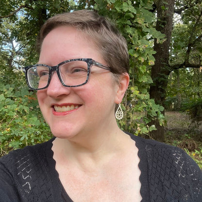 Person wearing glasses and a black sweater outdoors with greenery in the background. She is wearing sterling silver and white wood teardrop earrings with geometric snowflake-like designs